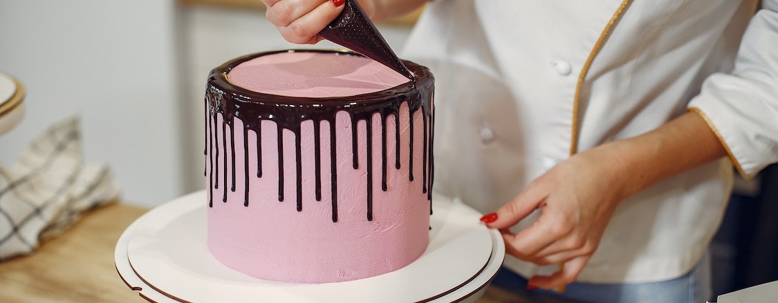 Chef piping chocolate on the edges of a pink round cake for a dripping effect on the sides
